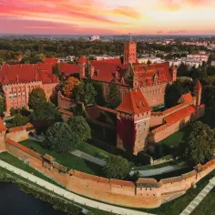 Potężny zamek w Malborku o czerwonych dachach, otoczony murami, w blasku zachodzącego słońca. Ciekawostki o tym miejscu czekają na odkrycie.