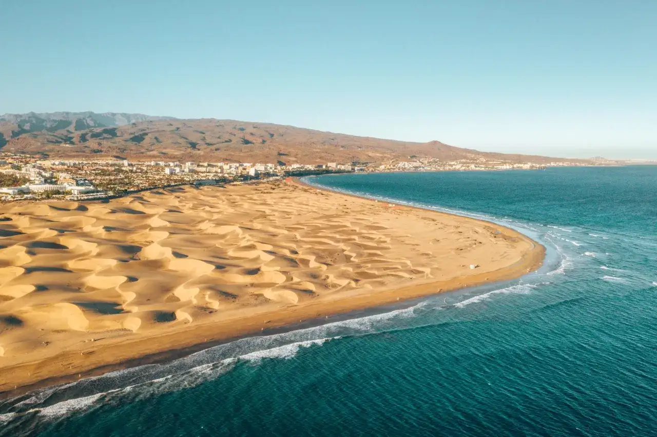 Piękne plaże Gran Canarii z wydmami i turkusowym oceanem. W tle widać góry i zabudowania miasta.