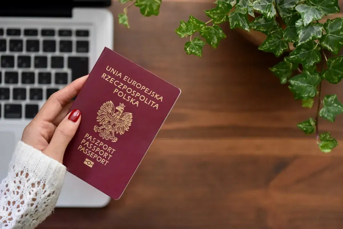 Dłoń trzyma polski paszport. Zastanawiasz się, czy emeryci mają zniżkę na paszport? Obok leży laptop i zielona roślina.