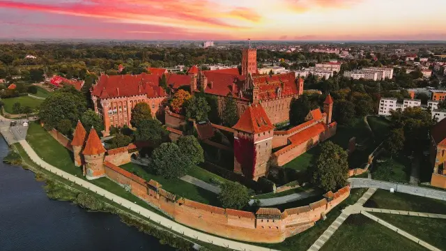 Potężny zamek w Malborku o czerwonych dachach, otoczony murami, w blasku zachodzącego słońca. Ciekawostki o tym miejscu czekają na odkrycie.
